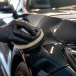 Dłoń w czarnej rękawiczce poleruje maskę czarnego samochodu za pomocą polerki mechanicznej
