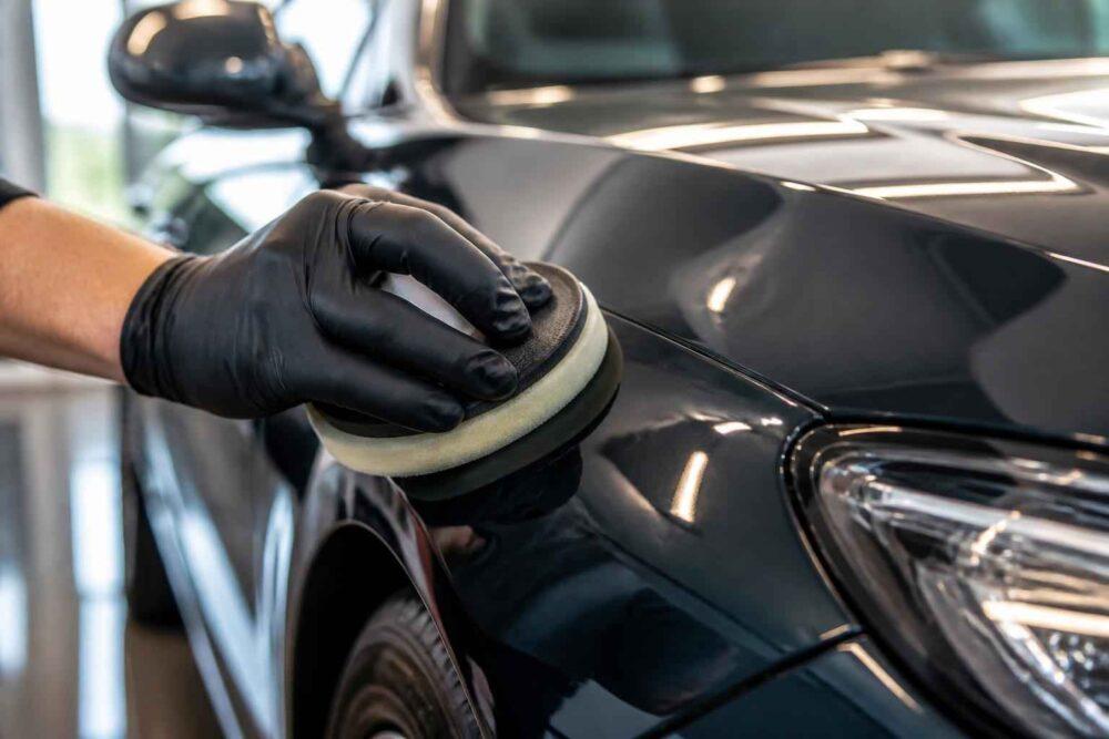 Dłoń w czarnej rękawiczce poleruje maskę czarnego samochodu za pomocą polerki mechanicznej